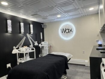Treatment room in a waxing salon with a padded bed, black blanket, multiple lamps and carts, and a lit 'Wax' logo on the wall.