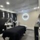 Treatment room in a waxing salon with a padded bed, black blanket, multiple lamps and carts, and a lit 'Wax' logo on the wall.