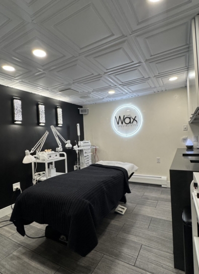 Treatment room in a waxing salon with a padded bed, black blanket, multiple lamps and carts, and a lit 'Wax' logo on the wall.