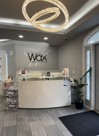 Reception area at The Wax Specialists with a curved white desk, logo on the gray wall, a ring-shaped chandelier, a potted plant, and brochure stands near the glass door.